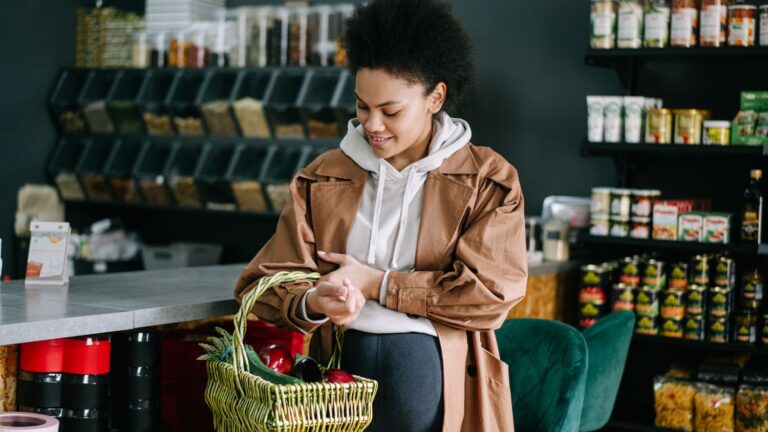 A pregnant woman in a beige coat over a white hoodie, carrying a basket of vegetables on her right arm.