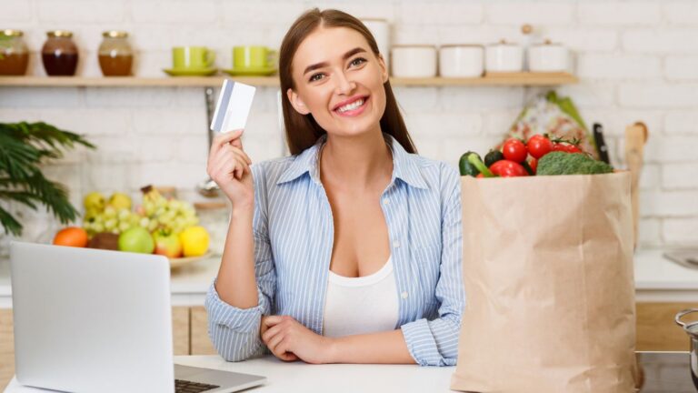 A smiling woman holding up a credit card while leaning on a kitchen countertop with a laptop and a bag of groceries in it.