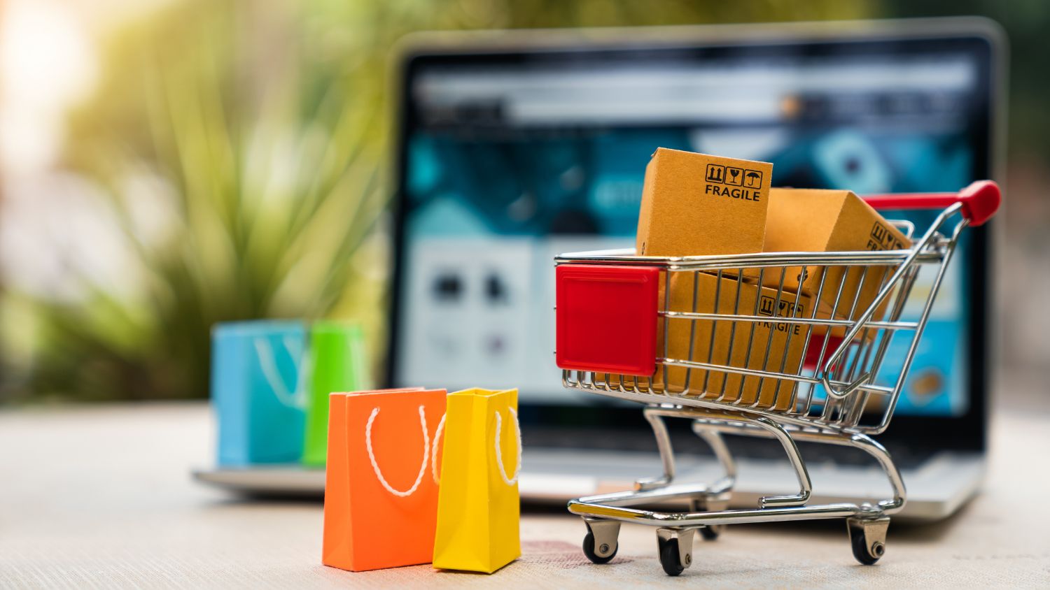 Focus shot on a small grocery cart with tiny packages in it. An open laptop can be seen in the background.