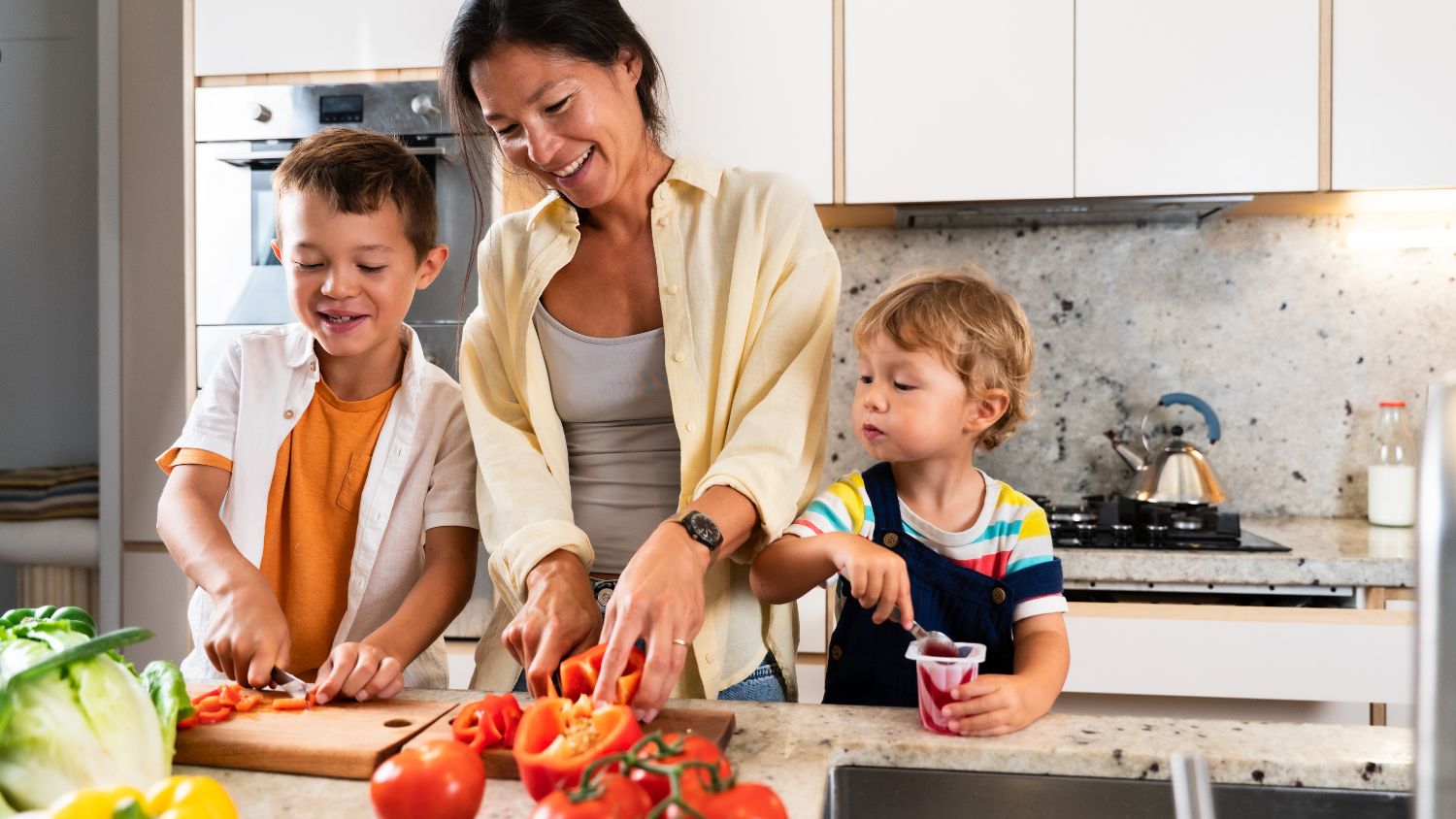 A mom cooking with her two young kids are standing on both sides on stools.