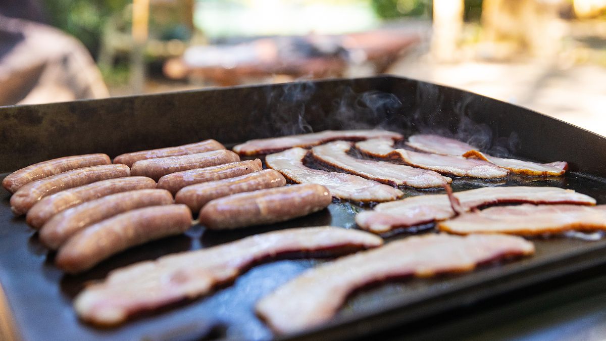 Eye level shot of an outdoor griddle cooking sausages and bacon in it.