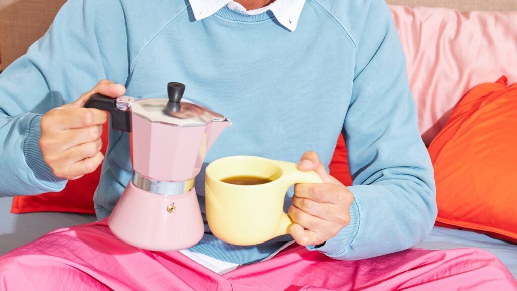 A person in a powder blue sweater with a white shirt underneath, and pink pants, holding an pink Moka pot on their right hand, and a powder yellow mug with coffee on the other.