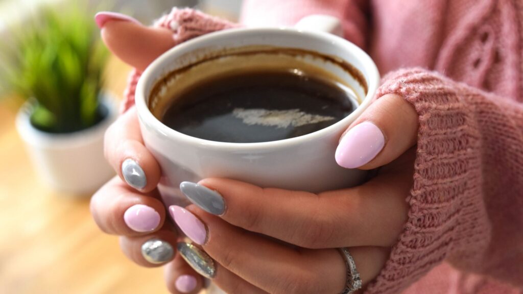 Focus shot on manicured hands holding a white cup with black coffee.