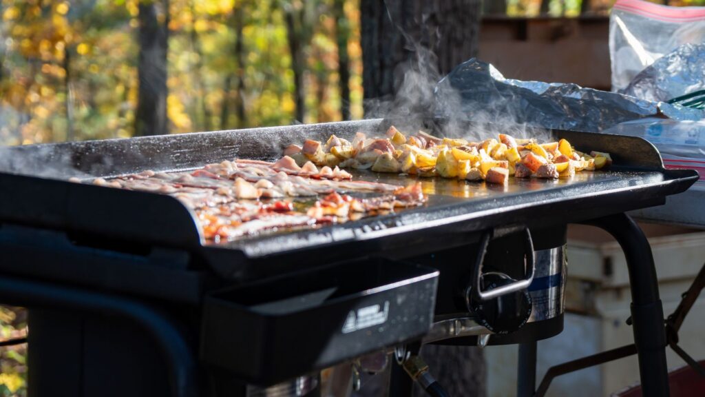 A griddle outdoors with bacon and potatoes cooking on it. Find out how to clean your outdoor griddle in this guide.