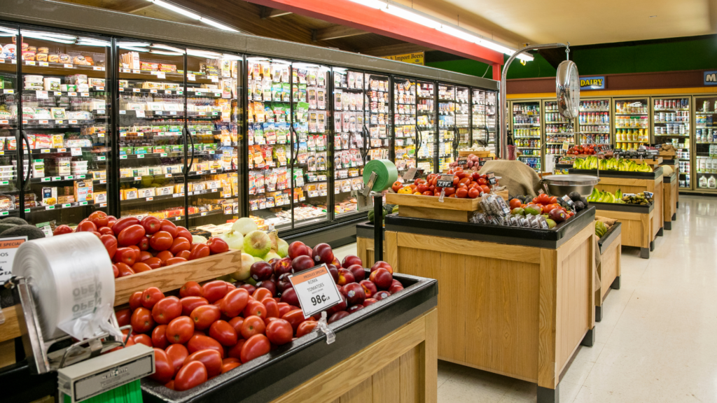A grocery store with fresh produce in wooden crate sections with frozen items and dairy in rows of fridge behind them.