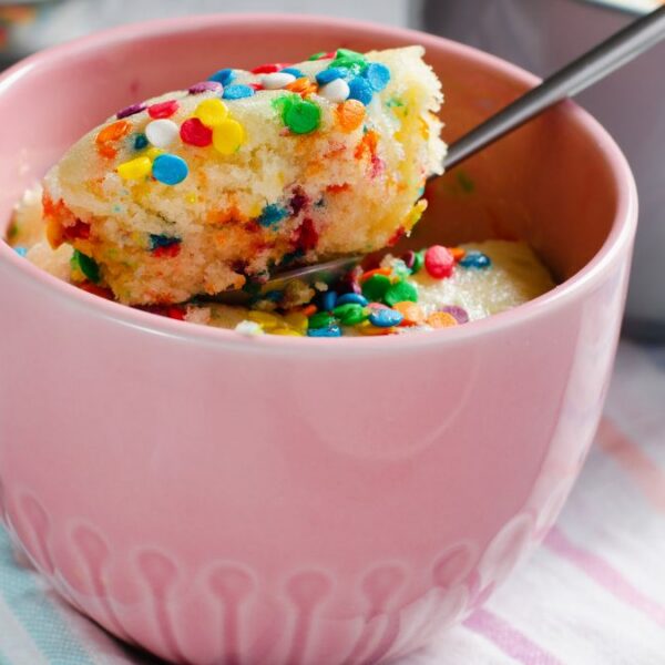 A mug cake with colorful sprinkles in a pink mug. A spoon is scooping out some of the cake.
