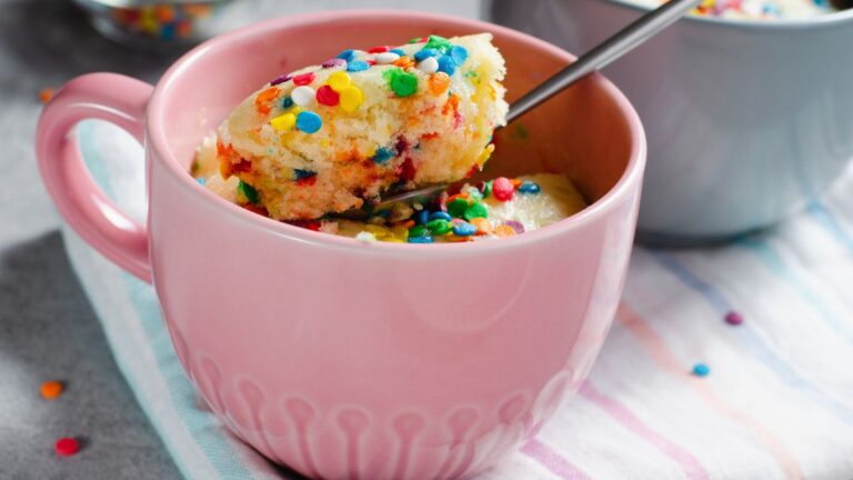 A mug cake with colorful sprinkles in a pink mug. A spoon is scooping out some of the cake.