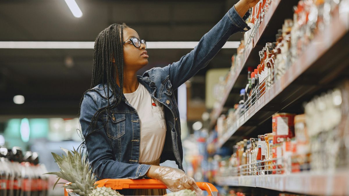 A woman buying groceries, reaching out for an item in the top shelf.