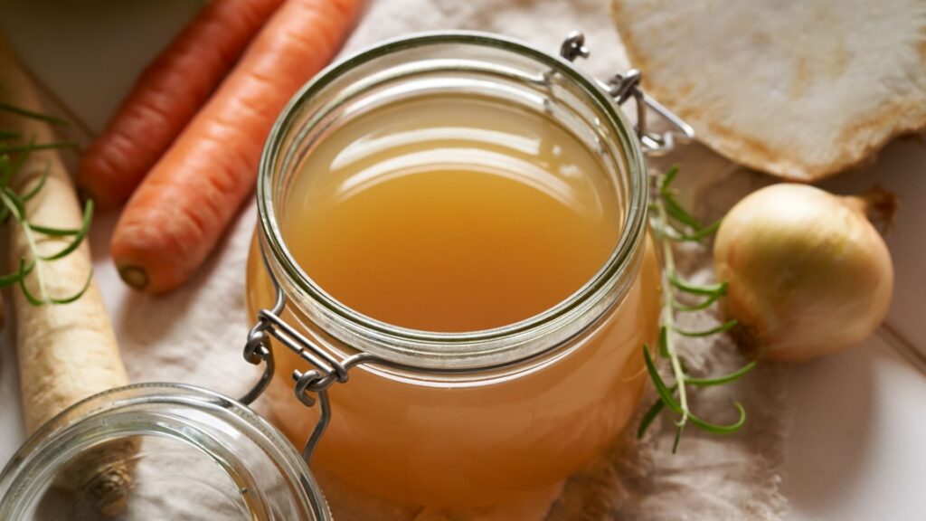 High-angle shot of a jar full of bone broth, surrounded by aromatics like onions, carrots and parsnips.