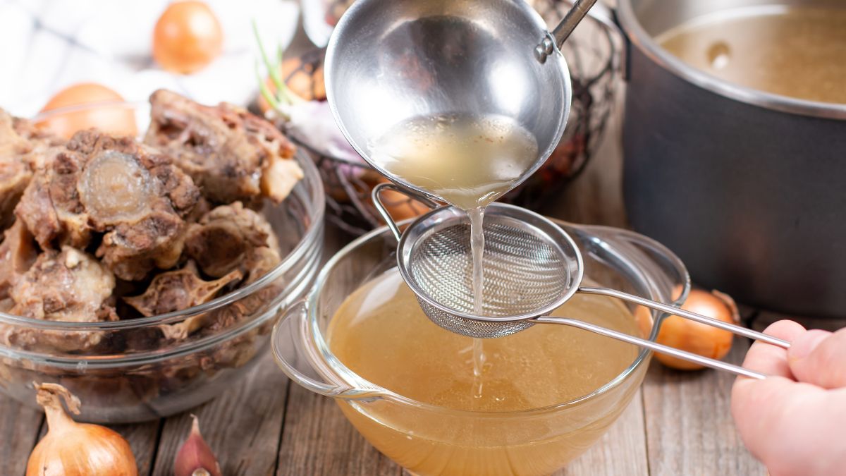 Hands straining the finished bone broth from a ladle to a clear bowl.