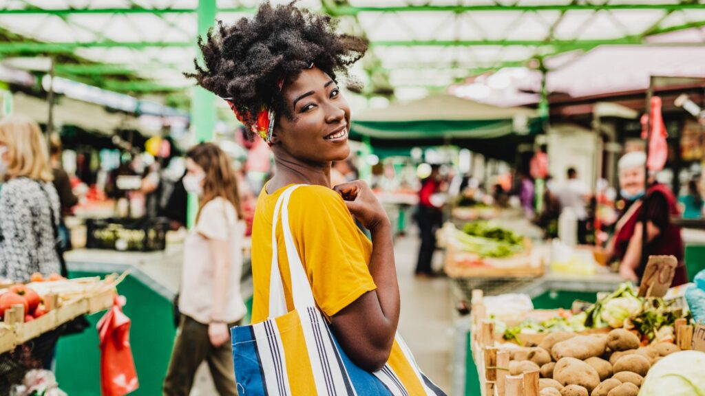 A woman in a yellow shirt carrying a shopping bag smiles as she walks along a farmers' market.