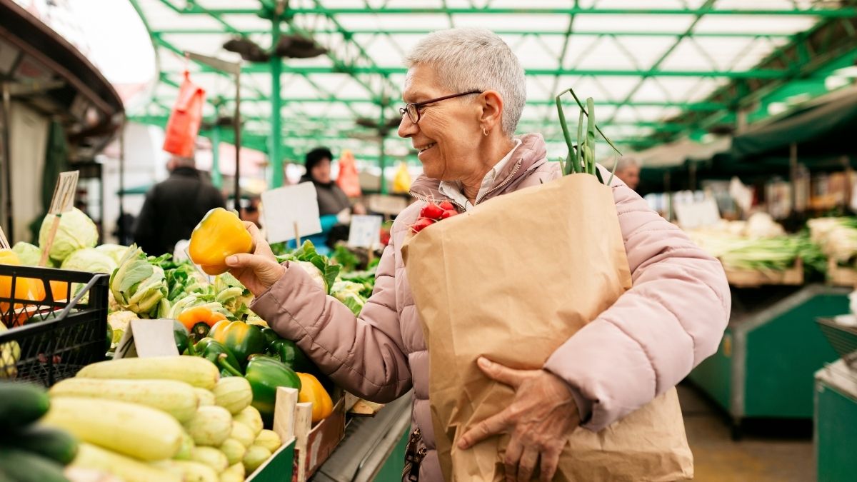 A woman happily shopping at a farmers market as she carries a paper bag with produce.