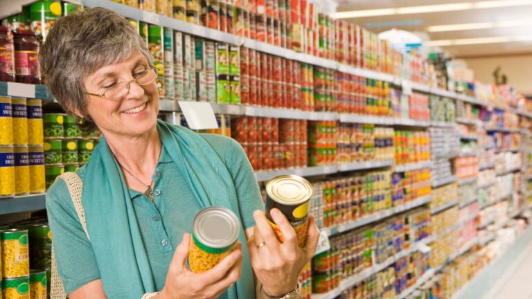 A woman comparing canned goods at a grocery store aisle. Here's Why Fall Is the Best Time to Stock Up on Canned Goods.