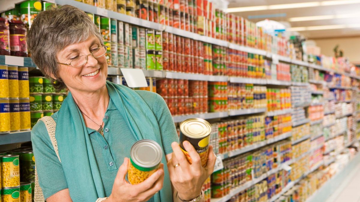 A woman comparing canned goods at a grocery store aisle. Here's Why Fall Is the Best Time to Stock Up on Canned Goods.