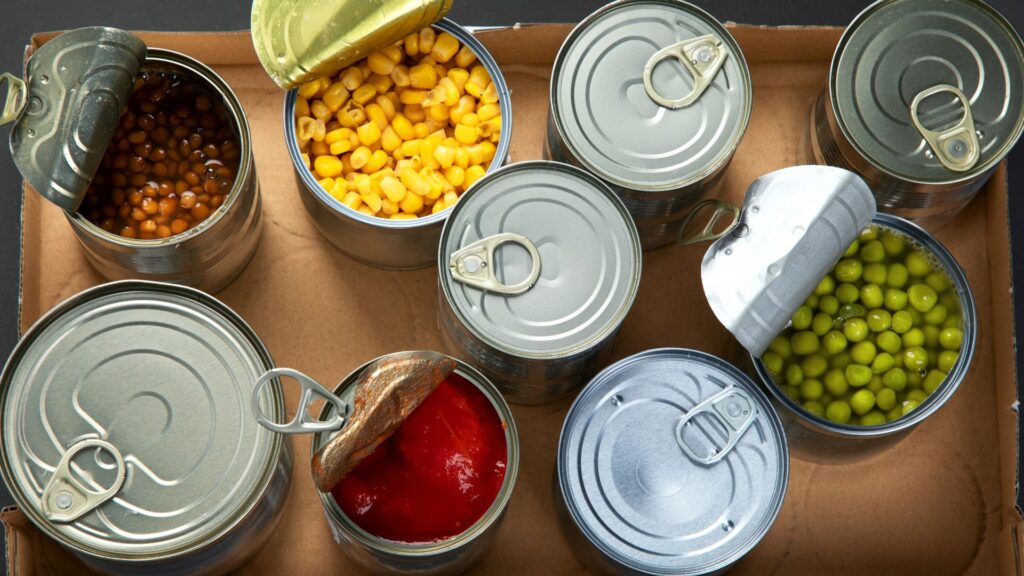 Flat lay shot of canned goods on a carton crate, some of them are opened and reveals corn, peas, tomato sauce, and lentils.