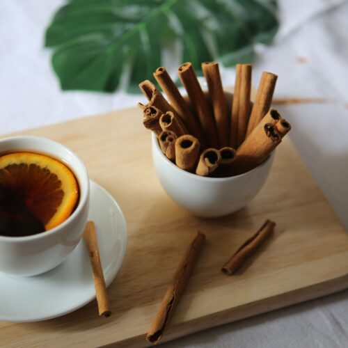 Spiced mulled apple cider in a white cup with an orange slice, beside a bowl of cinnamon sticks
