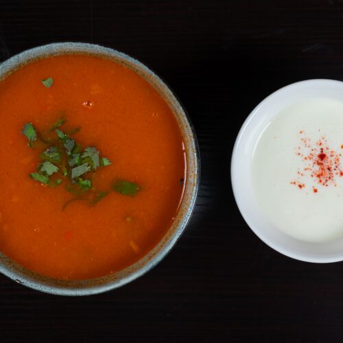 A bowl of tomato soup garnished with cilantro, served alongside yogurt and paprika.
