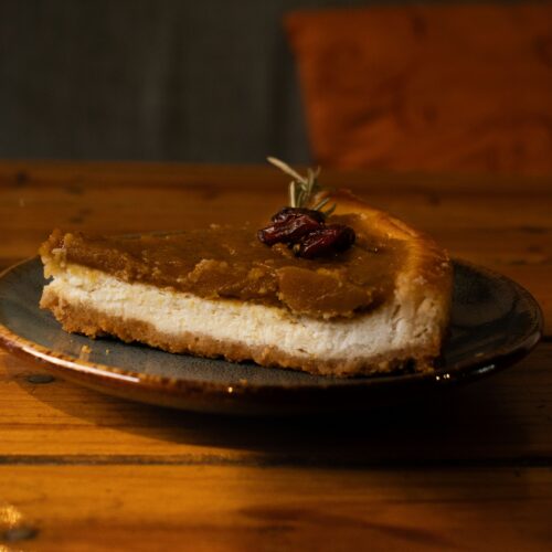 A slice of pumpkin cheesecake on a graham cracker crust, topped with cranberries and served on a dark plate