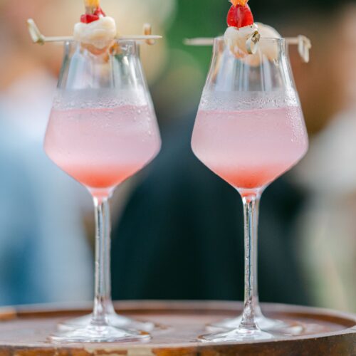 Two pink lychee cocktails garnished with whole lychees, served on a wooden tray