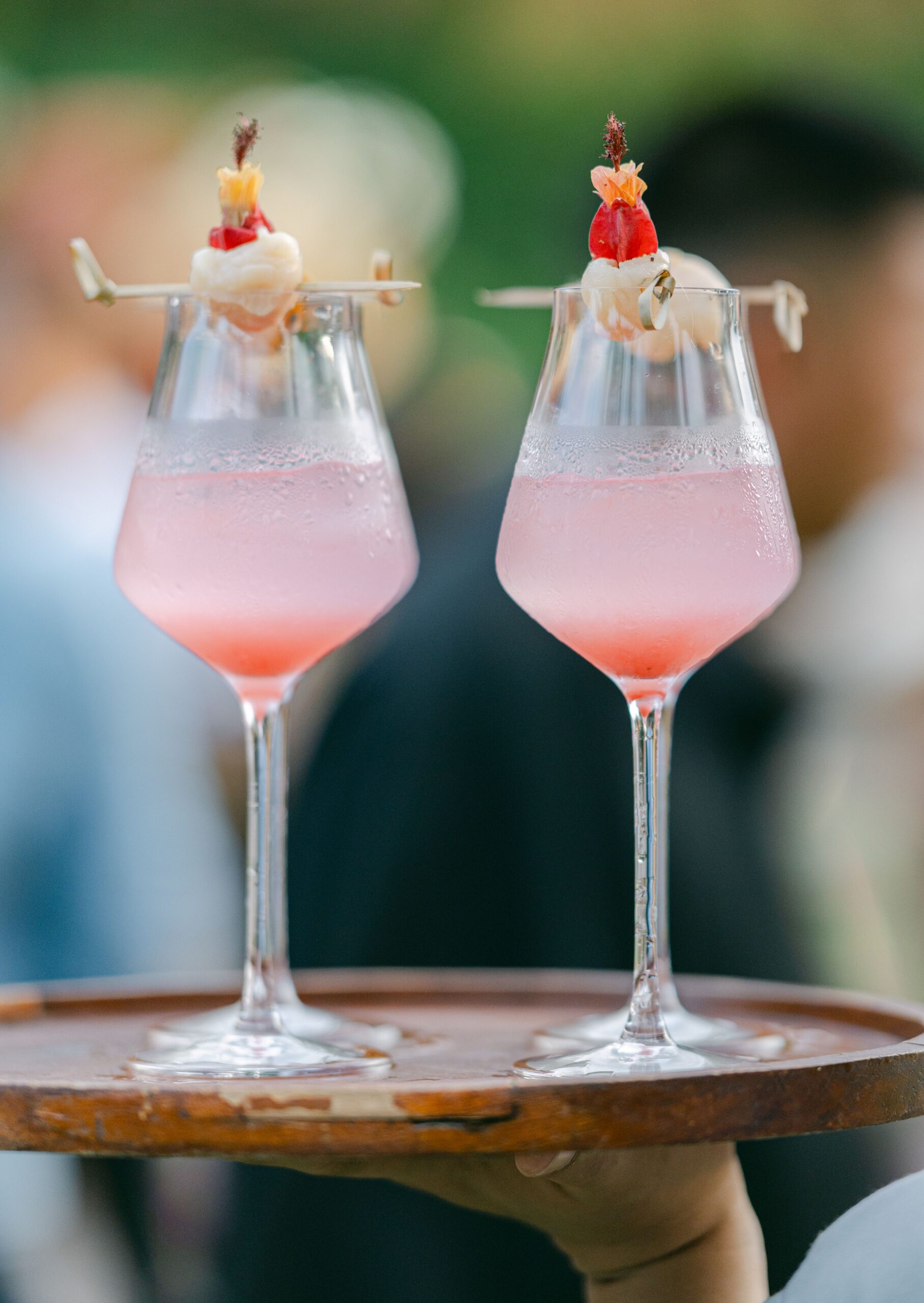Two pink lychee cocktails garnished with whole lychees, served on a wooden tray