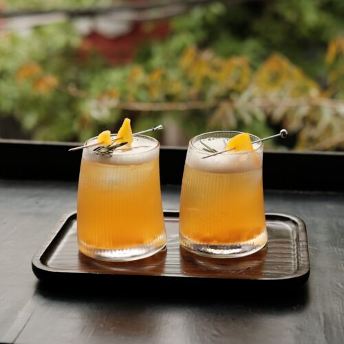 Two peach-colored bourbon cocktails garnished with citrus peel on a wooden tray outdoors