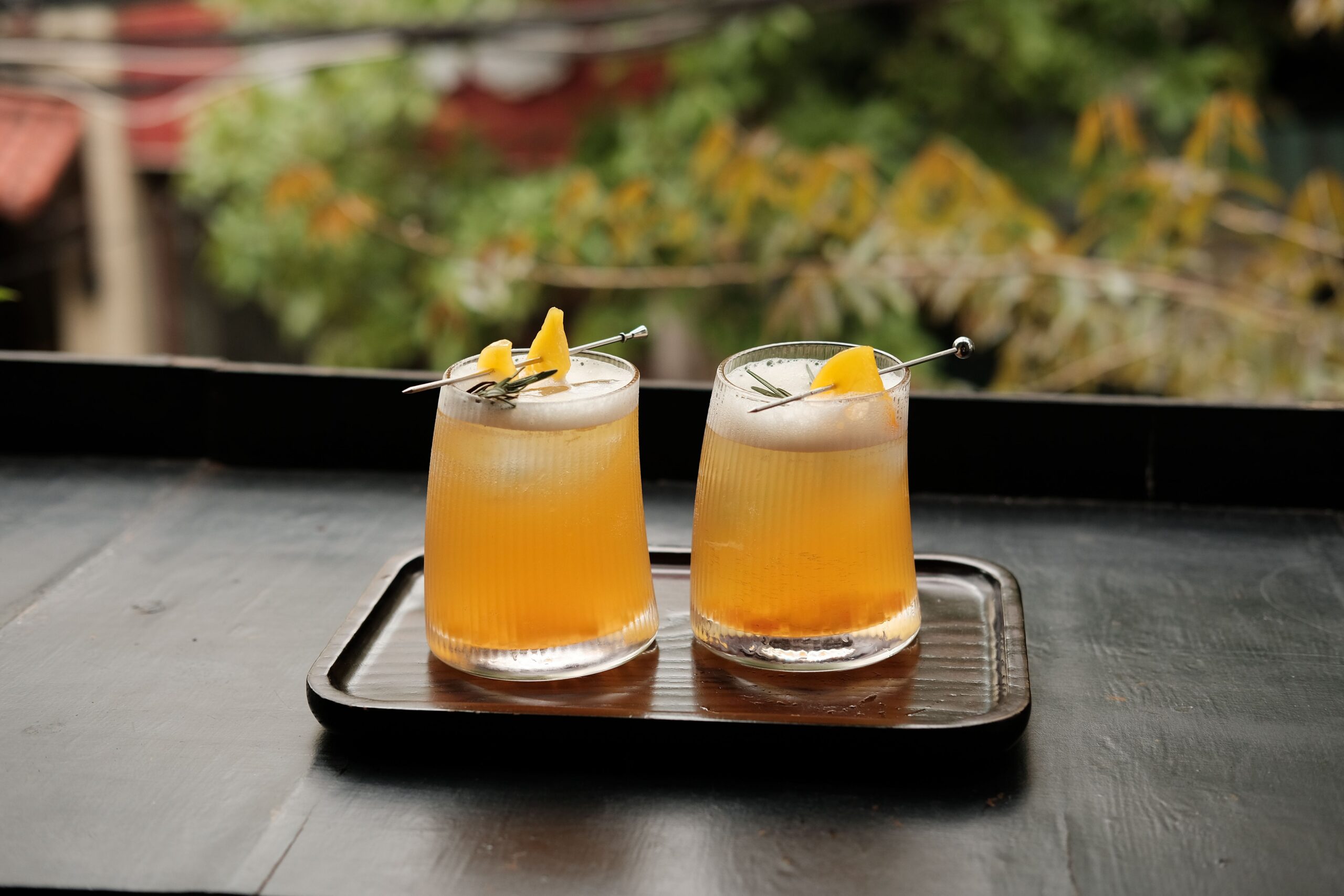 Two peach-colored bourbon cocktails garnished with citrus peel on a wooden tray outdoors
