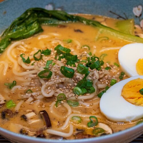 Close-up of a savory ramen noodle soup with eggs, greens, and sesame seeds.