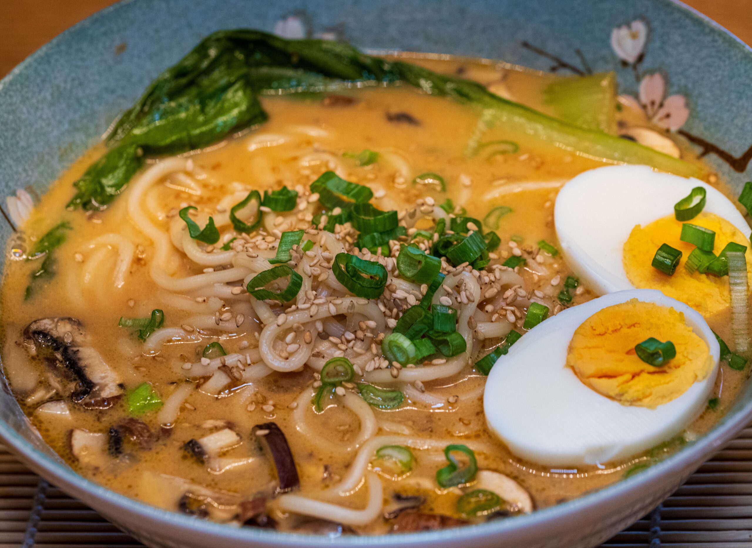 Close-up of a savory ramen noodle soup with eggs, greens, and sesame seeds.