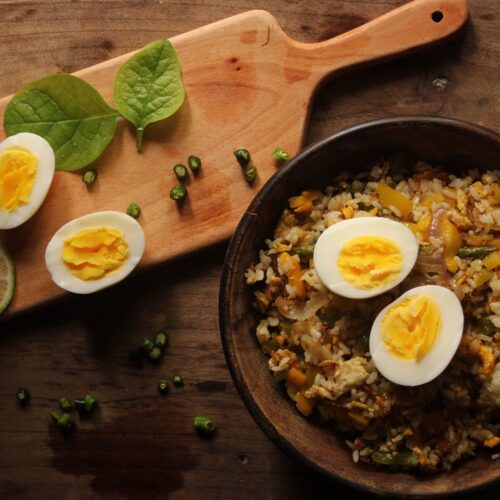 Top-down view of a bowl of fried rice with eggs and greens on a wooden board.