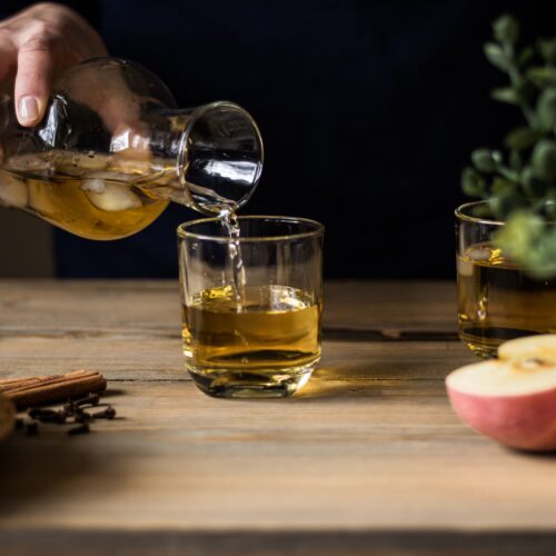 Apple cider being poured into glasses on a wooden table with apples and whole spices for a bourbon cider punch