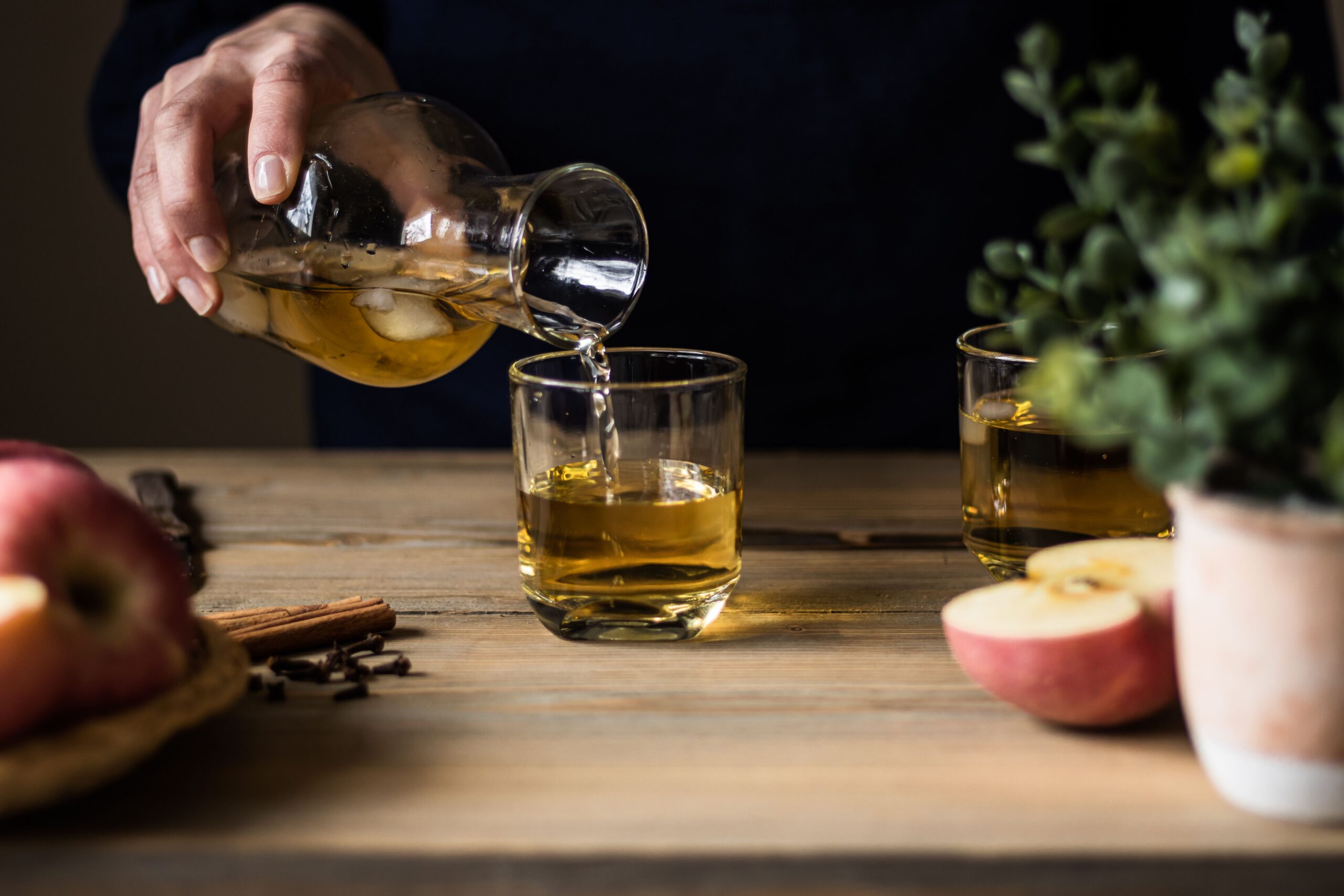 Apple cider being poured into glasses on a wooden table with apples and whole spices for a bourbon cider punch