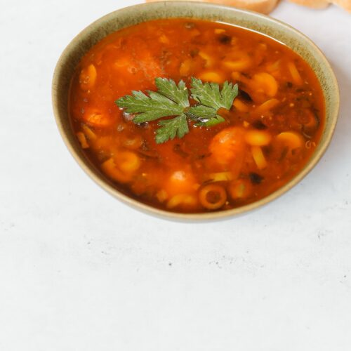 A bowl of minestrone soup with pasta, beans and vegetables in tomato broth, garnished with parsley and served with bread.