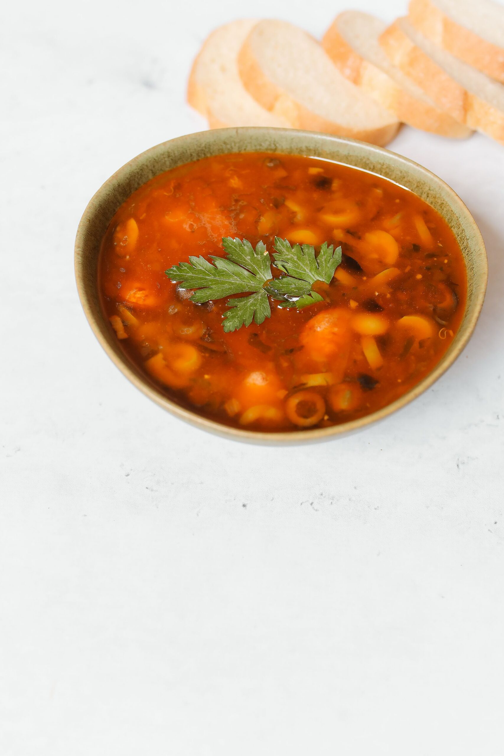A bowl of minestrone soup with pasta, beans and vegetables in tomato broth, garnished with parsley and served with bread.