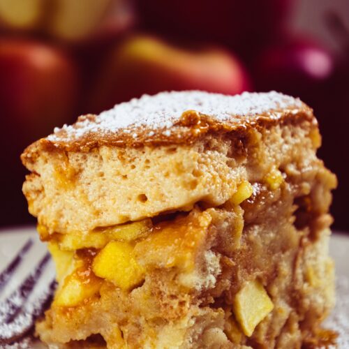 A square serving of apple cobbler with a soft golden baked topping over cinnamon-spiced apples, dusted with powdered sugar.