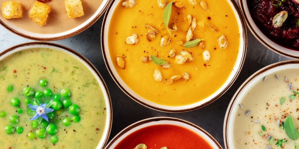 Flat lay shot of different bowls of soups, on a dark wood surface.