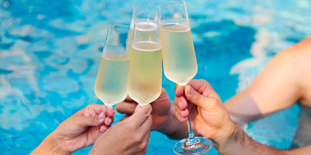 Four people's hands holding up a glass of champagne for a toast in a blue pool.
