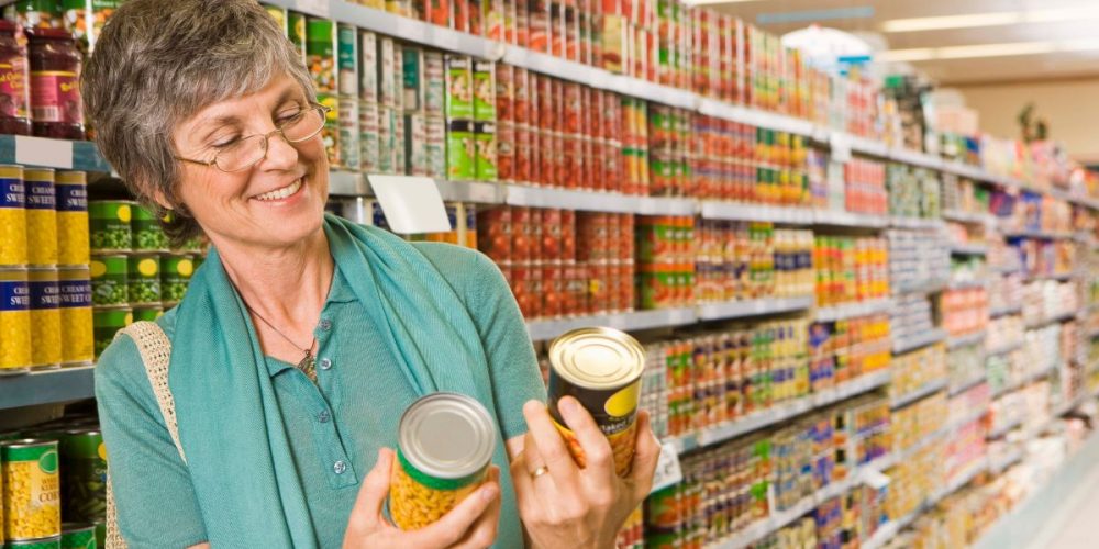 A woman comparing canned goods at a grocery store aisle. Here's Why Fall Is the Best Time to Stock Up on Canned Goods.