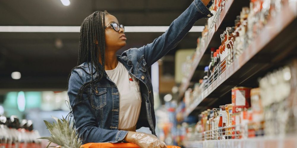 A woman buying groceries, reaching out for an item in the top shelf.