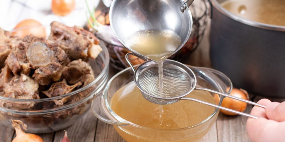 Hands straining the finished bone broth from a ladle to a clear bowl.