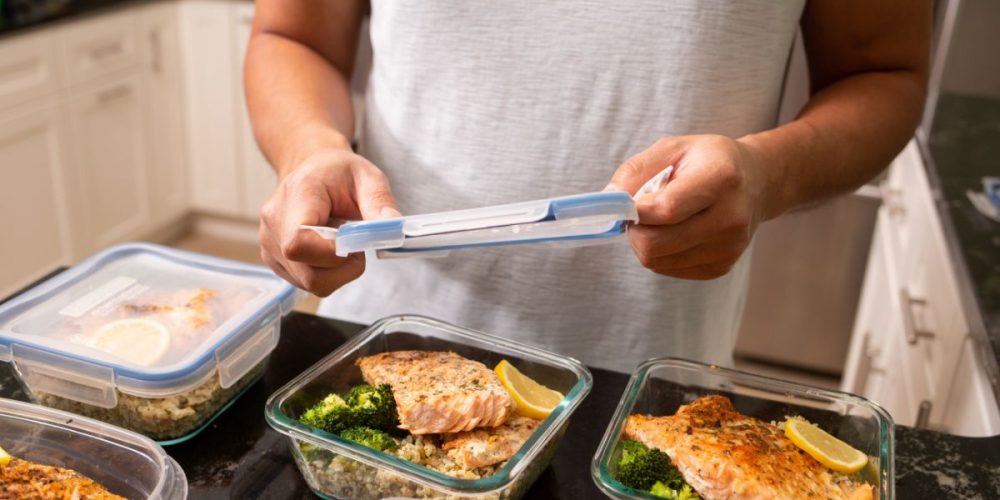 A person about to cover a container with meal prepped dish of salmon and vegetables. This article covers How to Meal Prep for Fall on a Budget