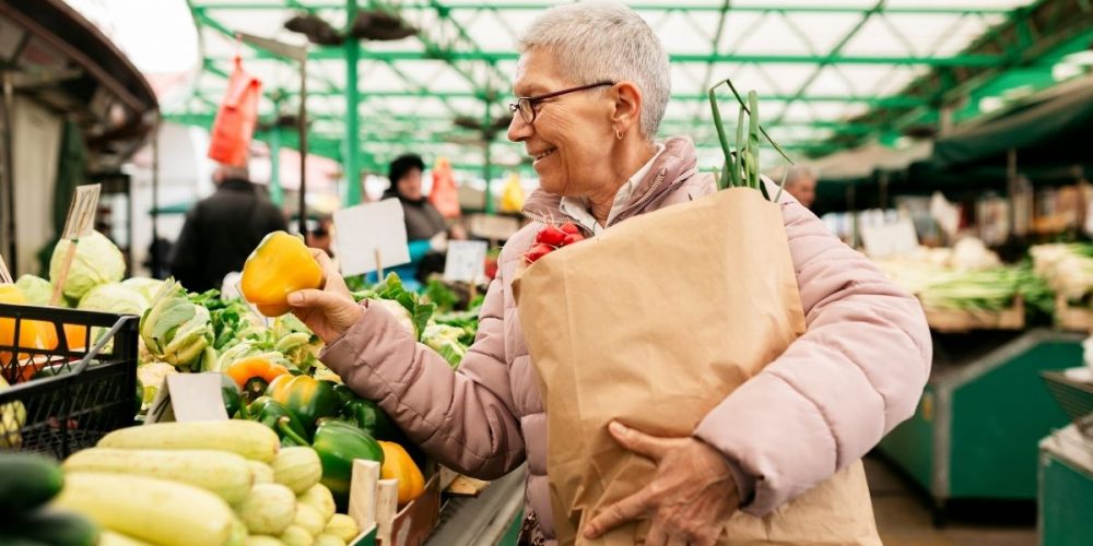 A woman happily shopping at a farmers market as she carries a paper bag with produce.