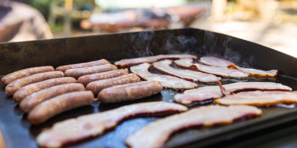 Eye level shot of an outdoor griddle cooking sausages and bacon in it.