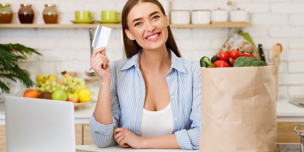 A smiling woman holding up a credit card while leaning on a kitchen countertop with a laptop and a bag of groceries in it.