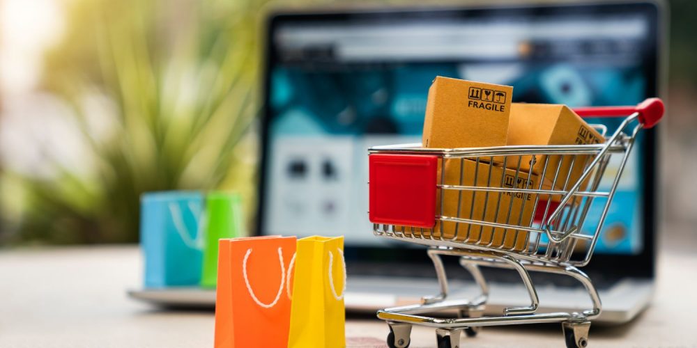Focus shot on a small grocery cart with tiny packages in it. An open laptop can be seen in the background.