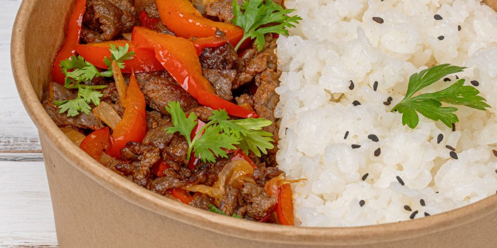 Close-up of a savory beef and vegetables dish with rice in a paper bowl.