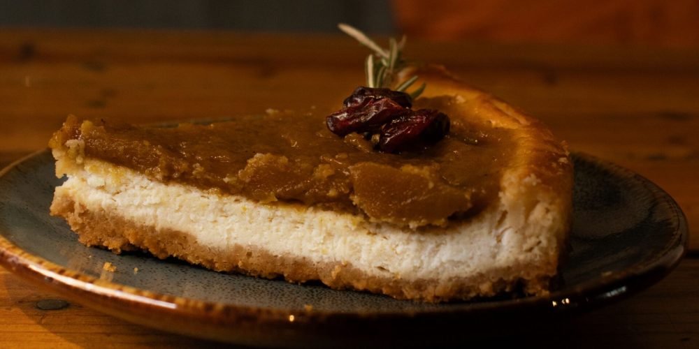 A slice of pumpkin cheesecake on a graham cracker crust, topped with cranberries and served on a dark plate
