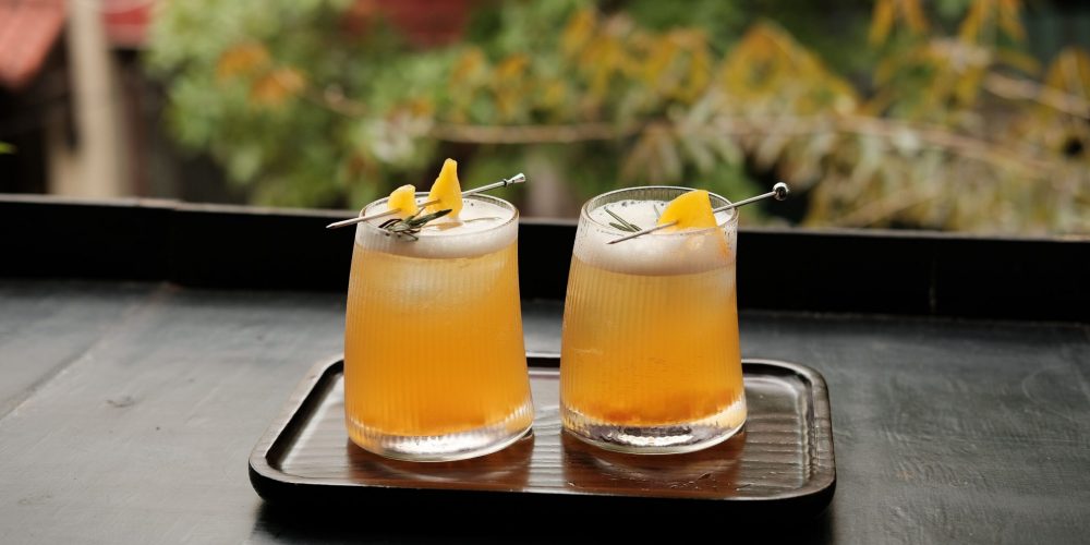 Two peach-colored bourbon cocktails garnished with citrus peel on a wooden tray outdoors