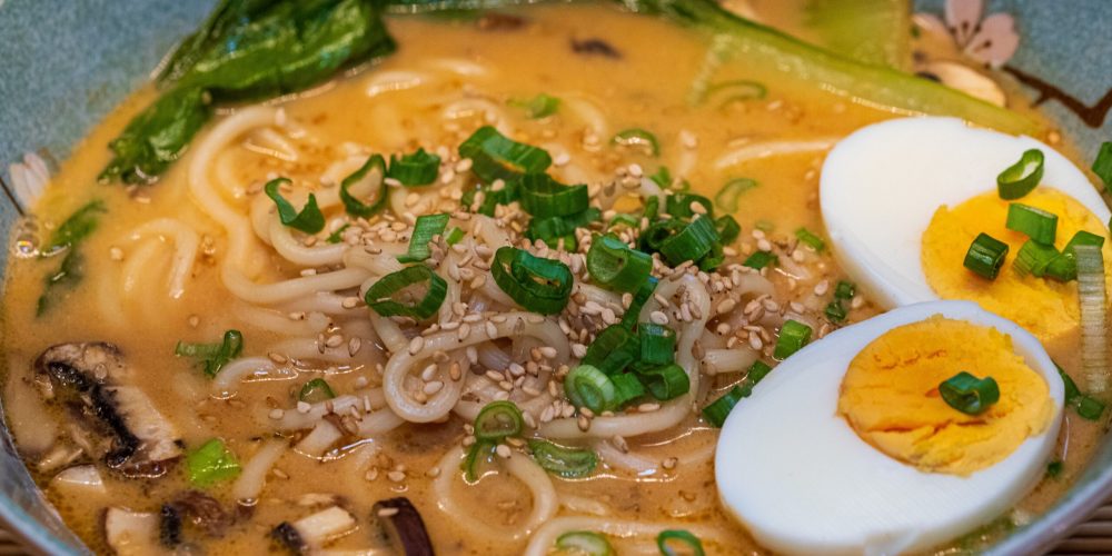 Close-up of a savory ramen noodle soup with eggs, greens, and sesame seeds.
