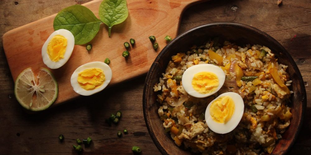Top-down view of a bowl of fried rice with eggs and greens on a wooden board.
