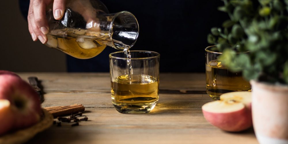 Apple cider being poured into glasses on a wooden table with apples and whole spices for a bourbon cider punch